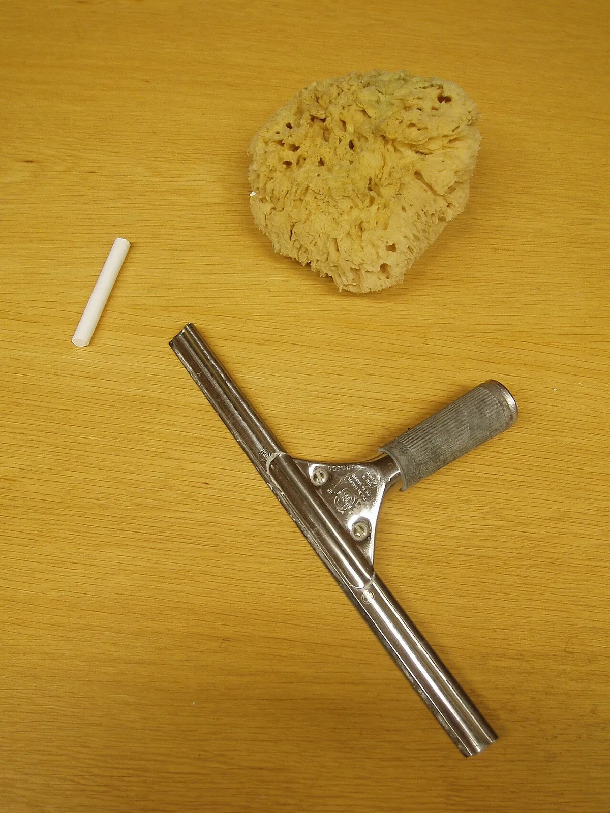 Een metalen raamwisser met rubberen strip naast een natuurspons en een stukje krijt op een houten tafel.